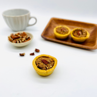  A 12-pack of mini Pecan Nut Pies displayed on a wooden tray, with one pie in the foreground and a small bowl of pecans beside a ceramic mug in the background.