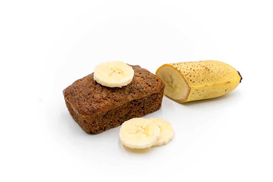 A delicious mini banana bread topped with fresh banana slices, displayed alongside a partially peeled banana on a white background. The image showcases the moist texture and rich color of the banana bread, highlighting its homemade appeal. Perfect for a bakery's promotional content.