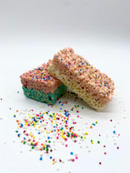 Two vibrant, multi-layered rice crispy treats are displayed, featuring distinct blue and pink sections topped with colorful sprinkles. The treats are placed on a white surface, with additional sprinkles scattered around, enhancing their festive appearance. These eye-catching, crunchy delights are perfect for adding a pop of color and fun to any dessert table.