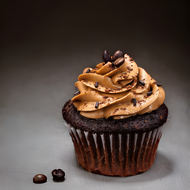 A decadent mocha cupcake featuring a rich, dark chocolate base topped with a generous swirl of mocha-flavored buttercream frosting. The frosting is elegantly decorated with chocolate sprinkles and a few whole coffee beans, adding a touch of sophistication. The cupcake sits on a dark, textured surface, highlighting its luxurious appearance. Perfect for showcasing a gourmet treat that combines the bold flavors of coffee and chocolate