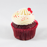  A delightful red velvet cupcake topped with smooth, creamy cream cheese frosting. The rich red color of the cupcake contrasts beautifully with the white frosting, which is elegantly swirled and adorned with small red sprinkles and a decorative red candy heart. The cupcake is presented in a matching red liner, making it a perfect treat for special occasions or celebrations.