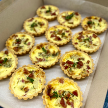 Load image into Gallery viewer, A box of 12 mini quiches topped with fresh herbs and crispy bacon, showcasing a golden crust and creamy filling.
