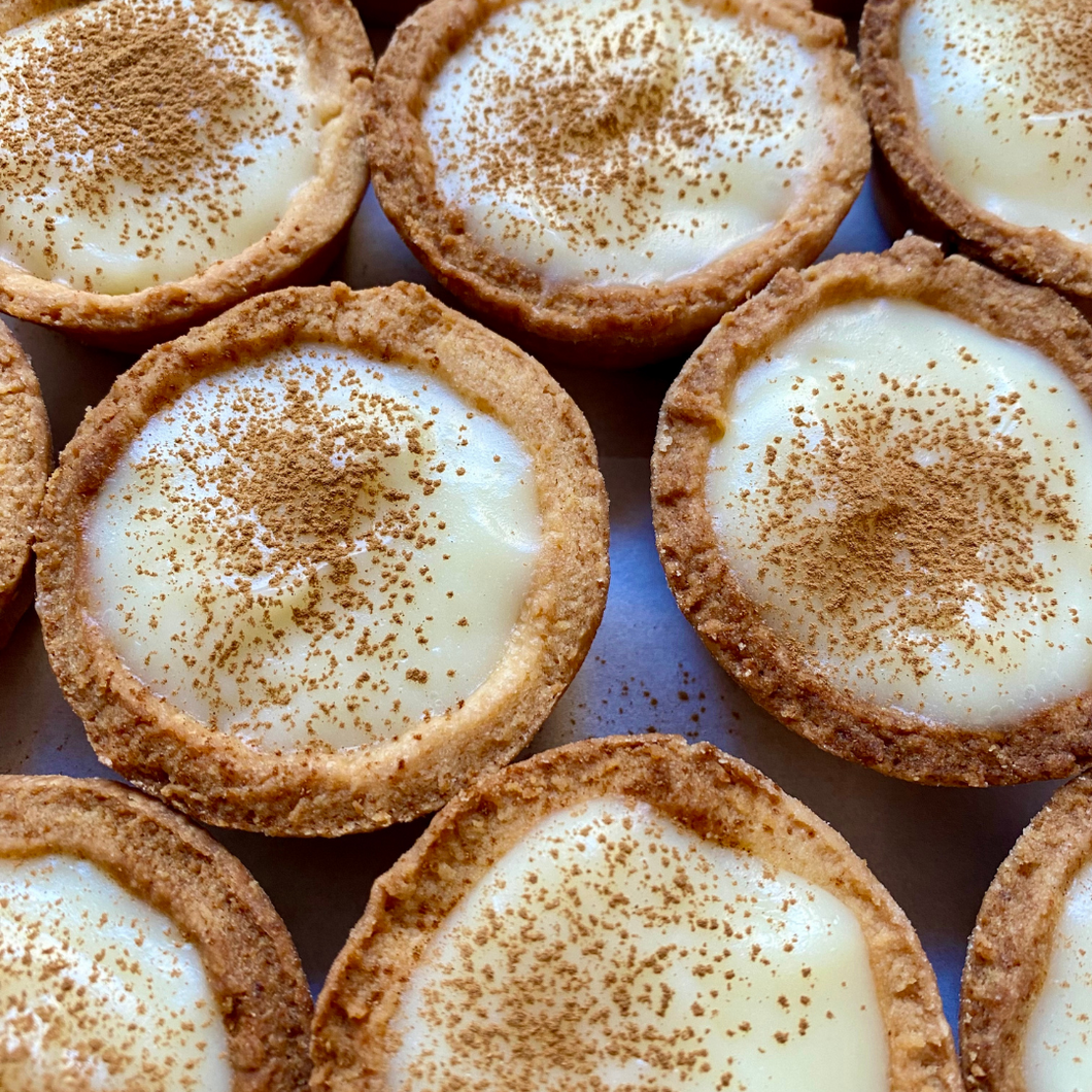 Close-up of 12 mini milk tarts with golden shortcrust pastry, topped with a light sprinkle of cinnamon, arranged neatly on a baking tray